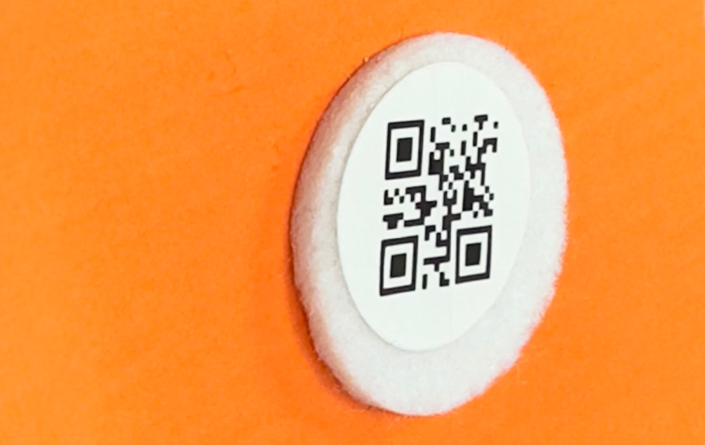 Bild på en rund etikett med QR-kod monterad på en något större möbeltass, fäst på kartongunderlag.