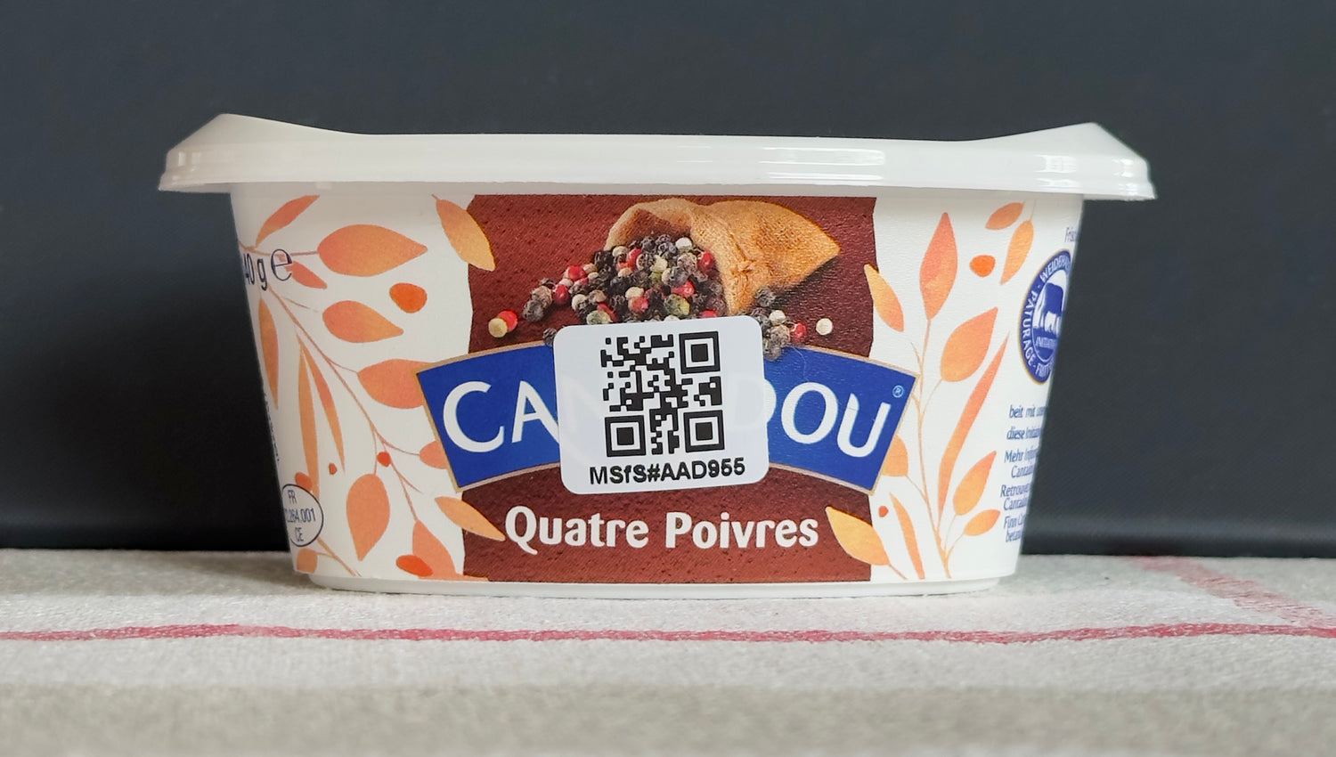 Bild på en fyrkantig vit etikett med QR-kod fäst på sidan av en plastförpackning med lock innehållande färskost.